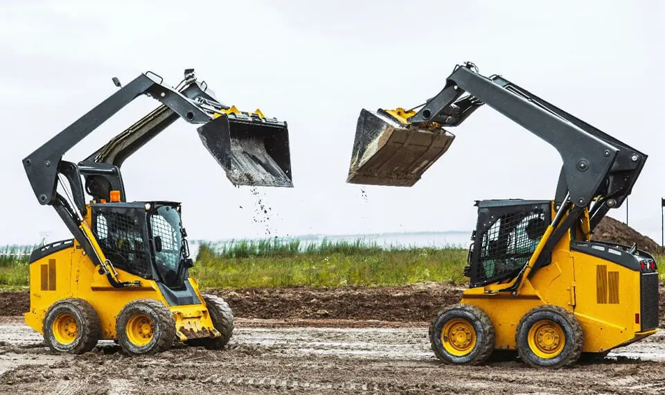 Two skid steers on a job site with their buckets elevated in the air.