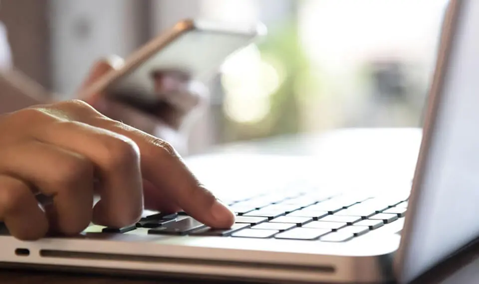 A photo of a person typing on their laptop computer while holding a mobile device in their other hand.